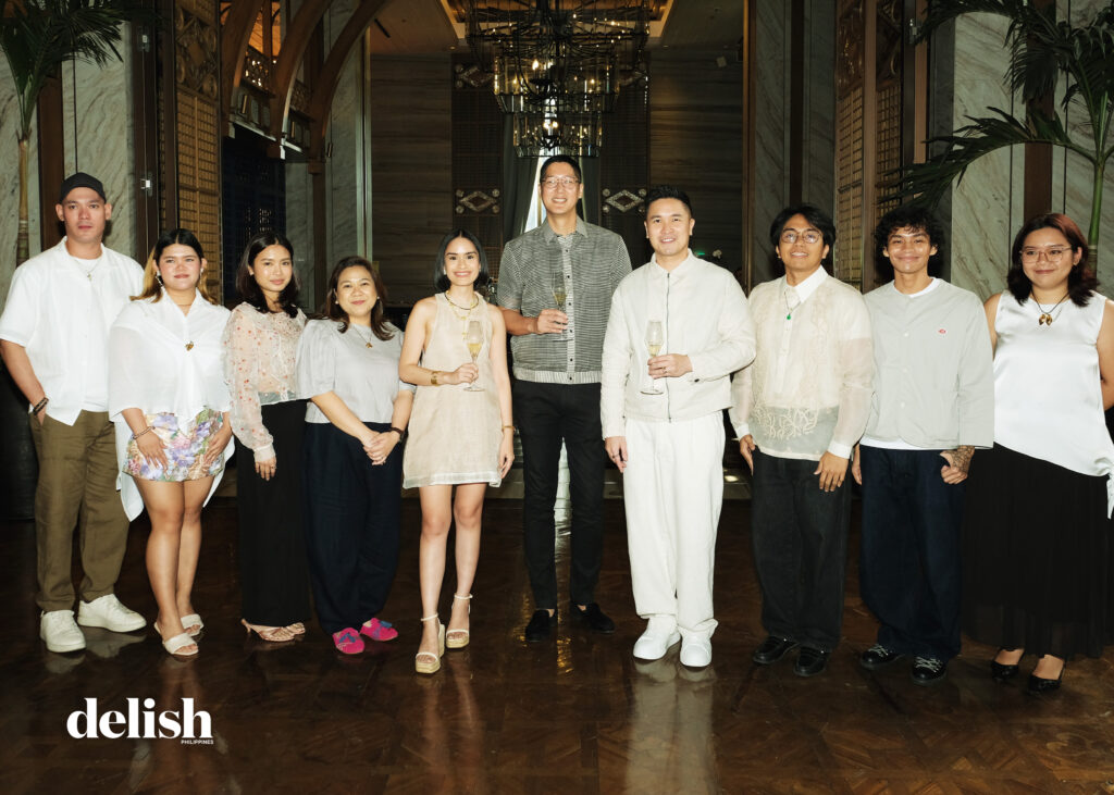 Group of eleven people in light, semi-formal outfits posing for a group photo at an upscale venue, several holding champagne flutes; Delish logo bottom-left.
