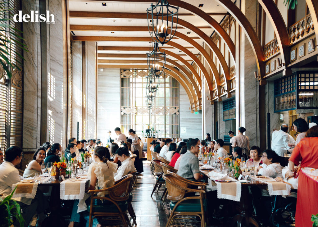 Elegant dining hall filled with guests at long white-clothed tables beneath curved wooden arches and chandeliers.
