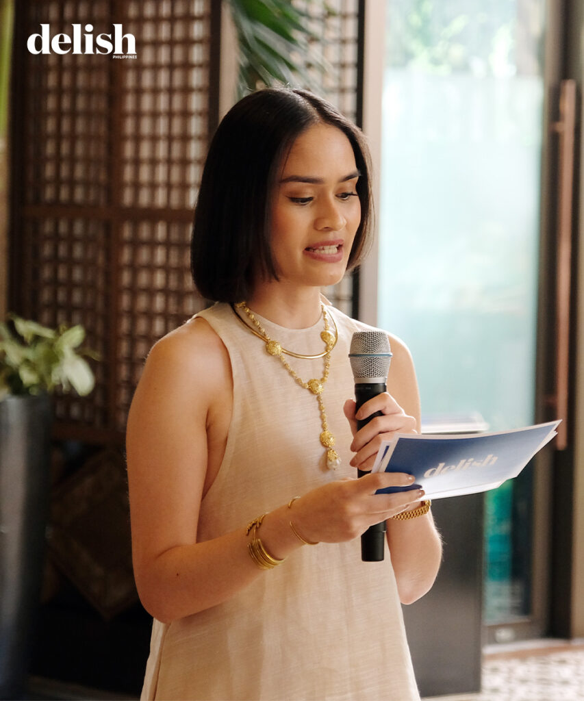 Woman in a sleeveless beige dress speaks into a handheld microphone while holding notes, with the Delish Philippines logo in the corner.