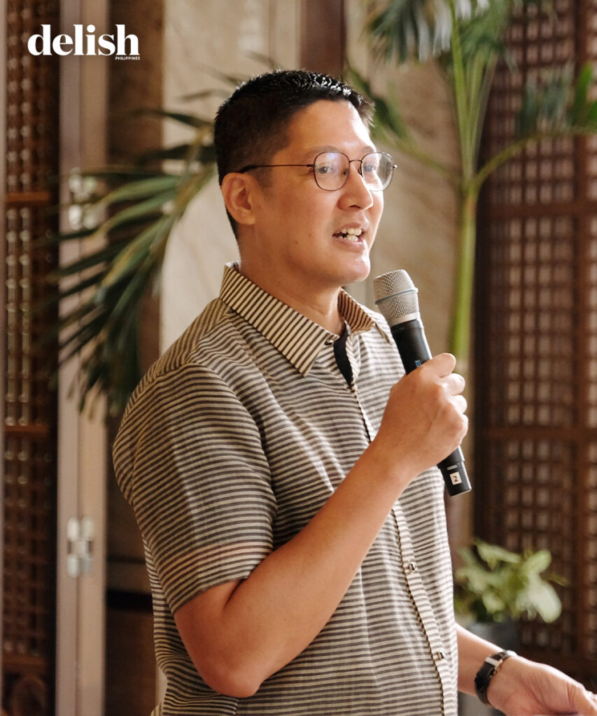 Man wearing glasses and a striped polo shirt speaks into a handheld microphone at an indoor event; delish Philippines logo visible in the corner