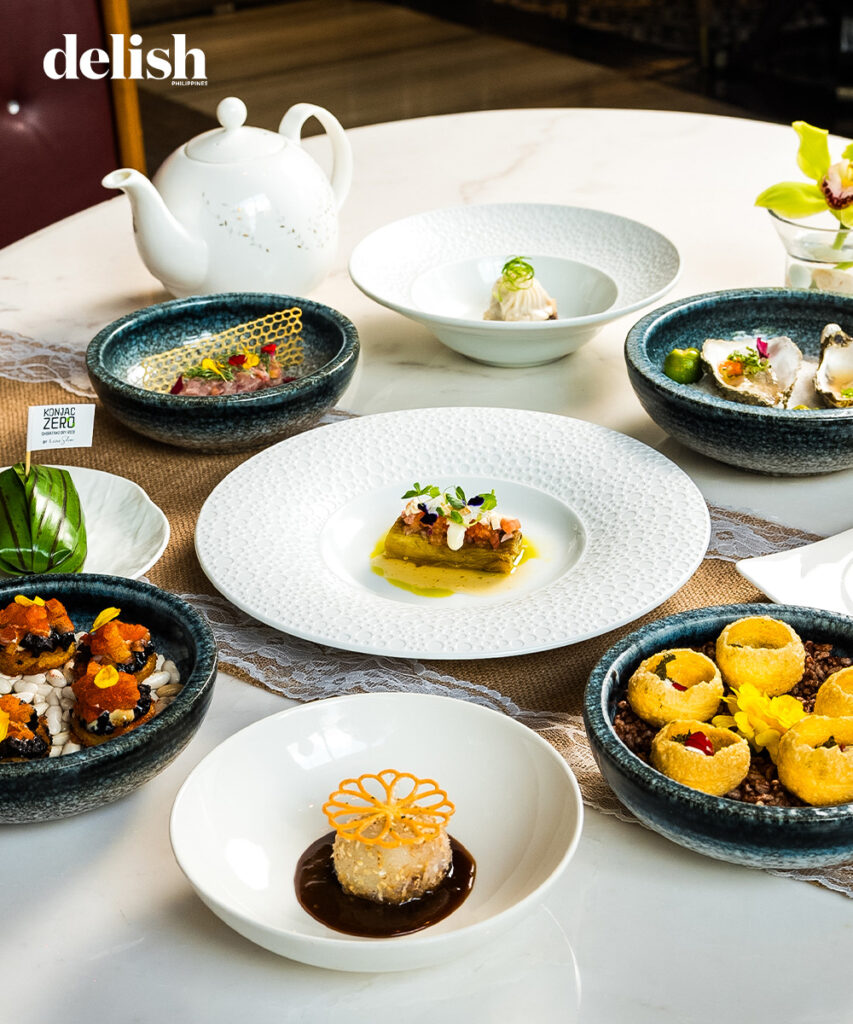 Gourmet dishes in blue bowls and white plates arranged on a marble table, with a white teapot and Delish Philippines logo in the corner.