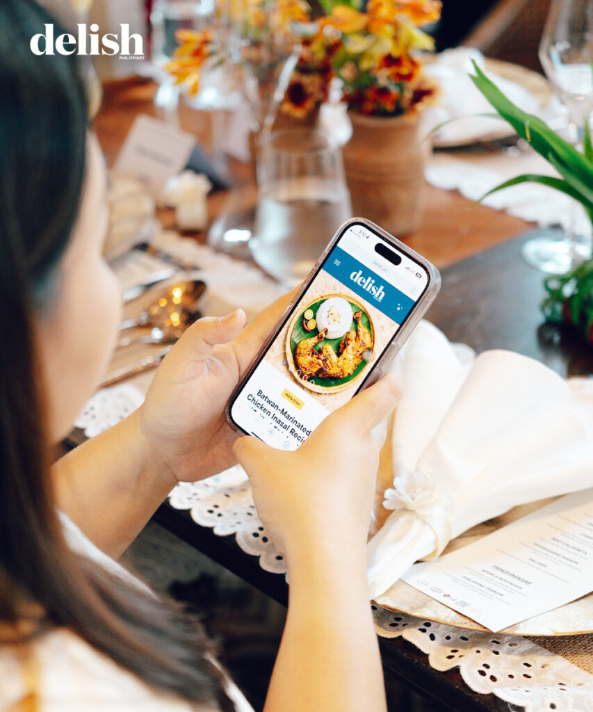 Person holding a smartphone showing a Delish Philippines recipe image at a dining table with utensils and flowers nearby.