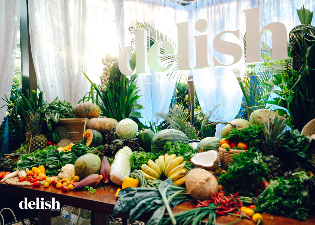 Abundant tropical fruits and vegetables arranged on a wooden table with a large 'delish' sign in the background.