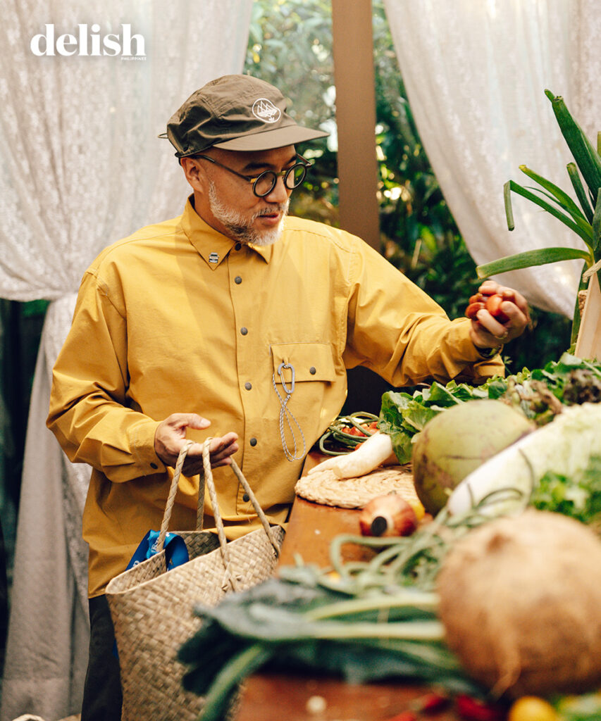 Older man with a gray beard and glasses, wearing a mustard shirt and cap, selecting produce at an outdoor market with a woven bag in hand.