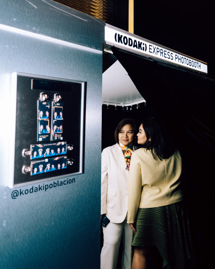 Two women posing at a Kodak Express photo booth, with photo strips visible inside the booth.