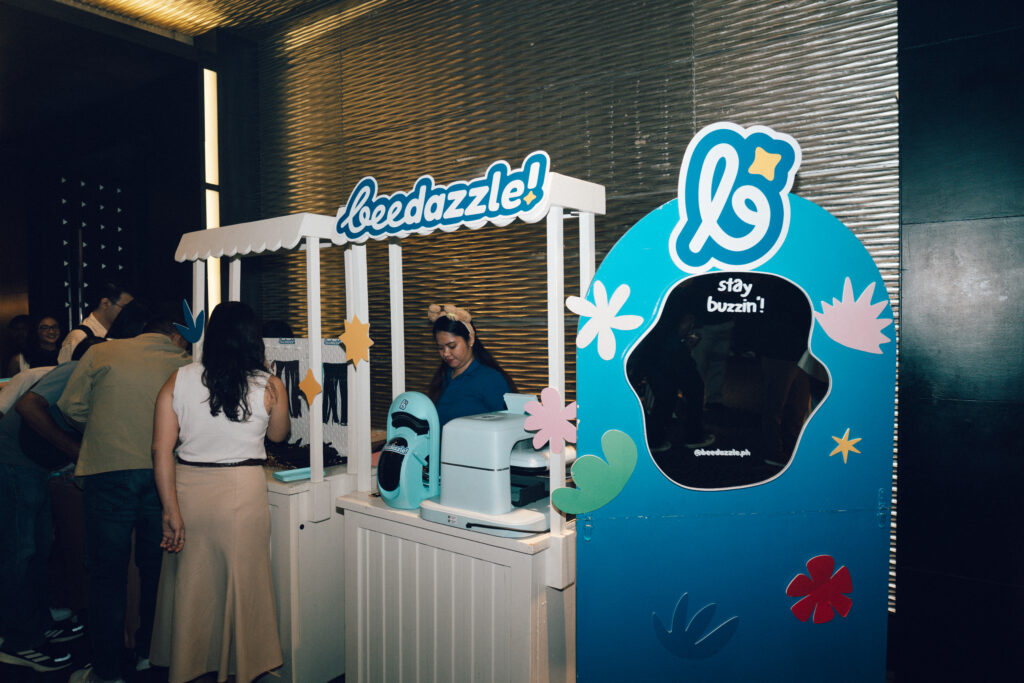 Promotional Beedazzle booth setup with a white canopy and attendees lined up indoors, turquoise photo-prop backdrop nearby.
