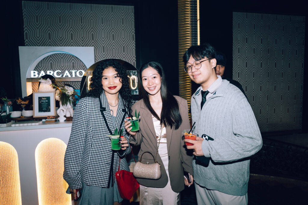 Three friends pose with colorful cocktails at a bar, with a 'BARCATO' sign in the background.