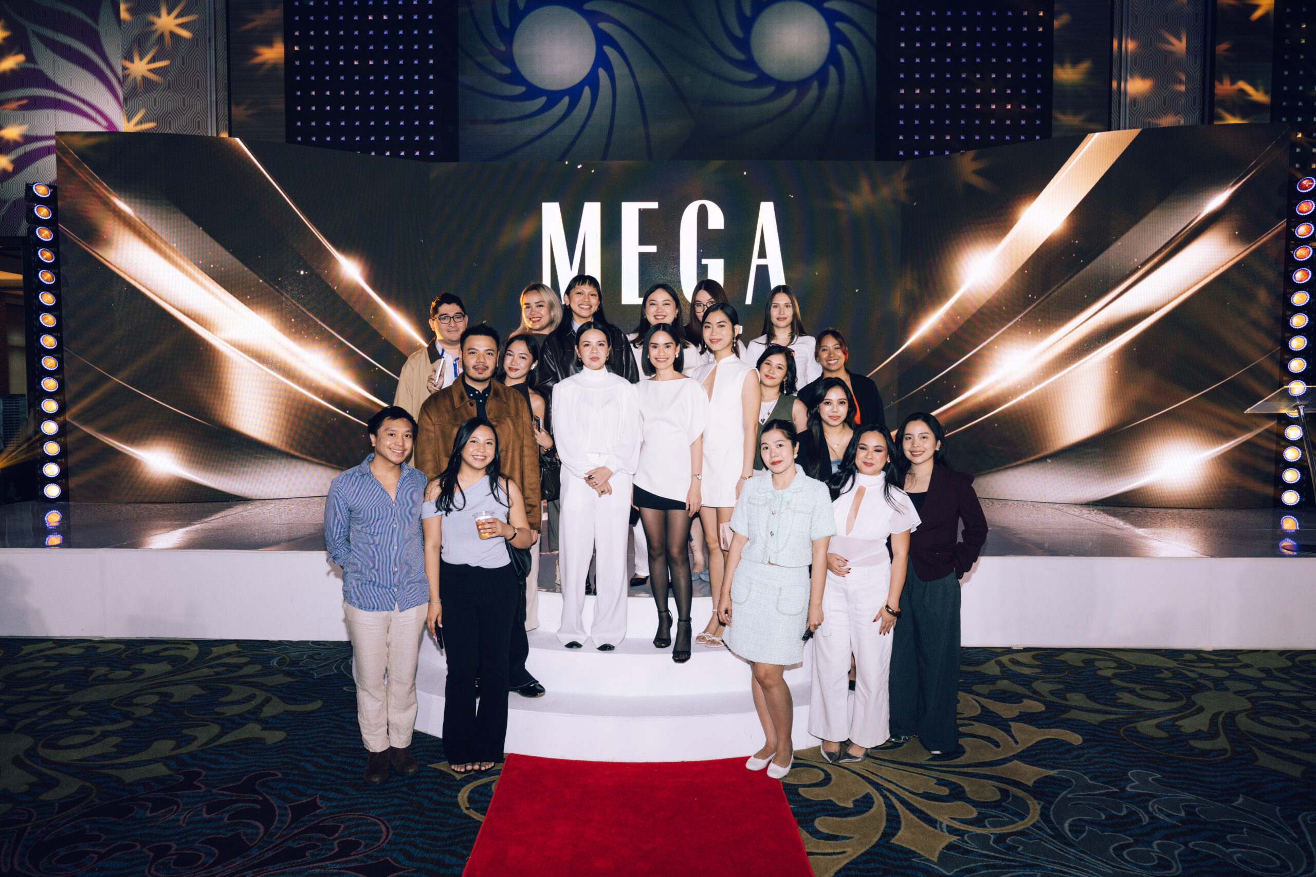 Group of people posing on a stage with a bold MEGA backdrop and gold lighting effects, red carpet in the foreground.
