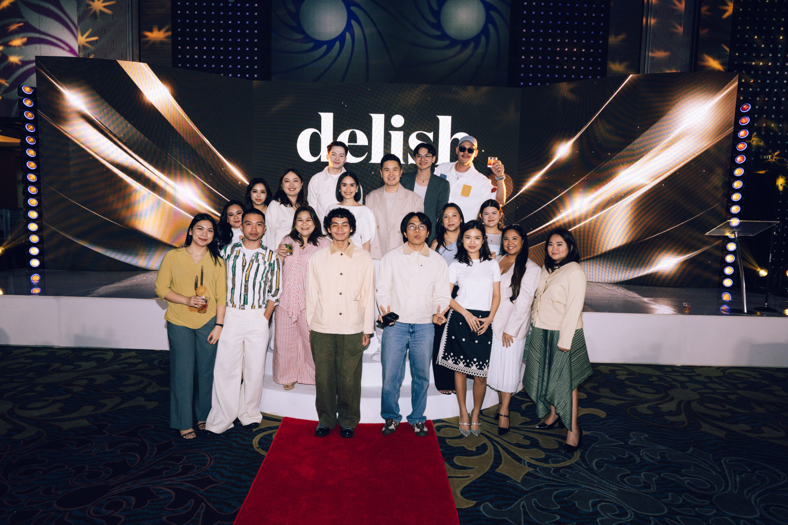 Group of about 16 people posing on a red carpet in front of a lit stage with a large 'delish' backdrop image behind them.