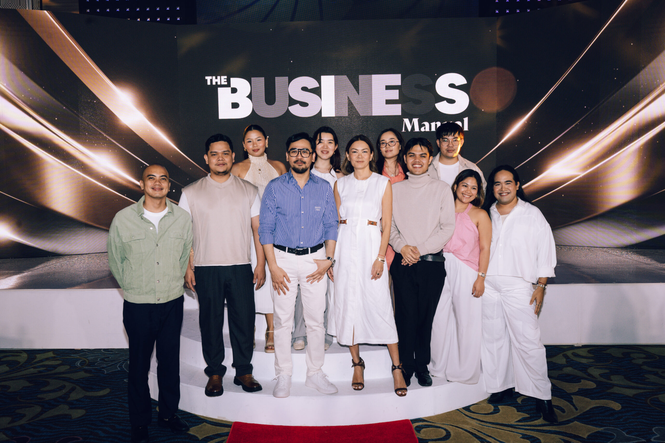 Group of diverse professionals posing on a stage with a large backdrop that reads 'THE BUSINESS' behind them.