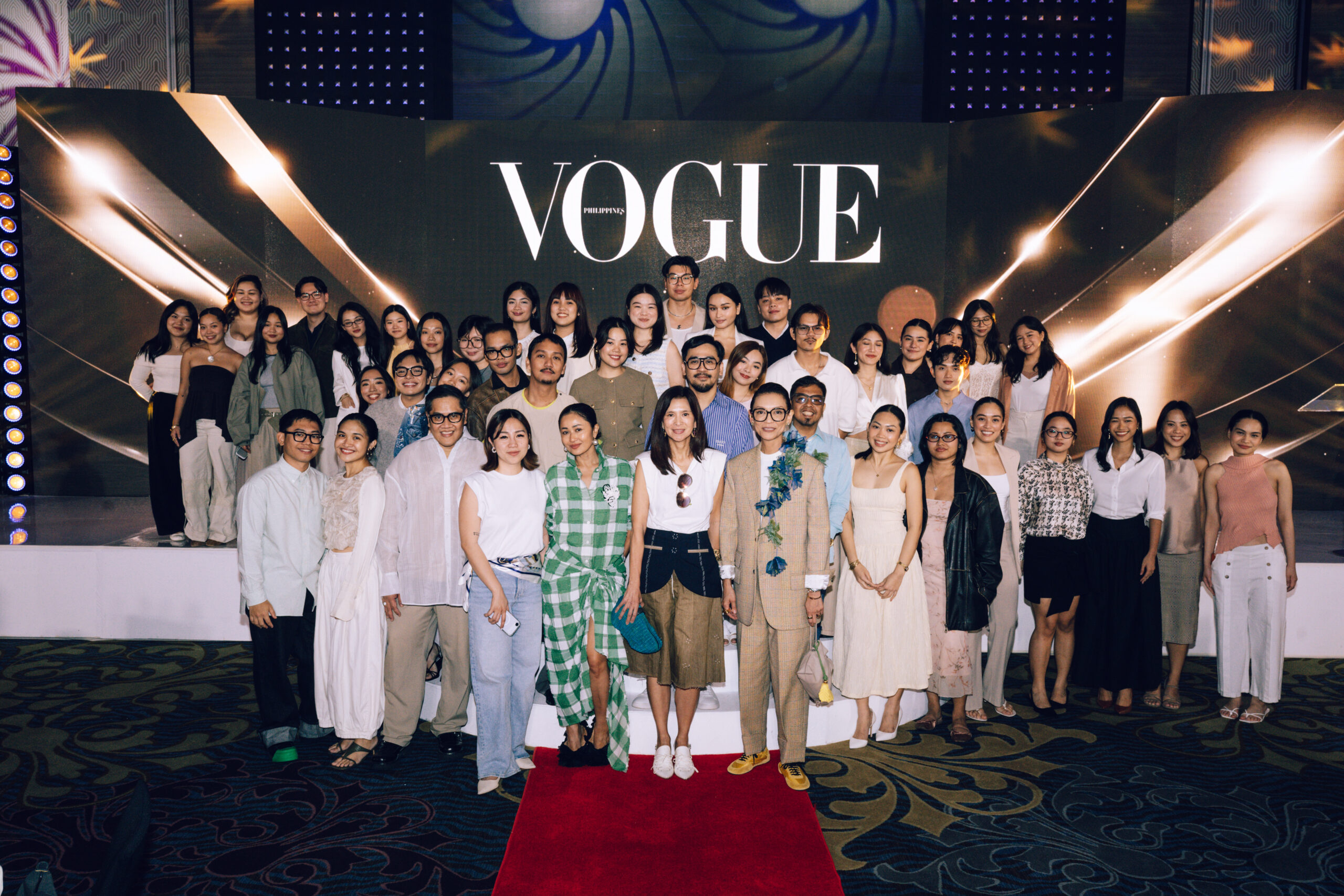 Group photo of fashion models and guests posing on a Vogue-branded stage with a red carpet and bright graphics in the background, smiling for the camera.