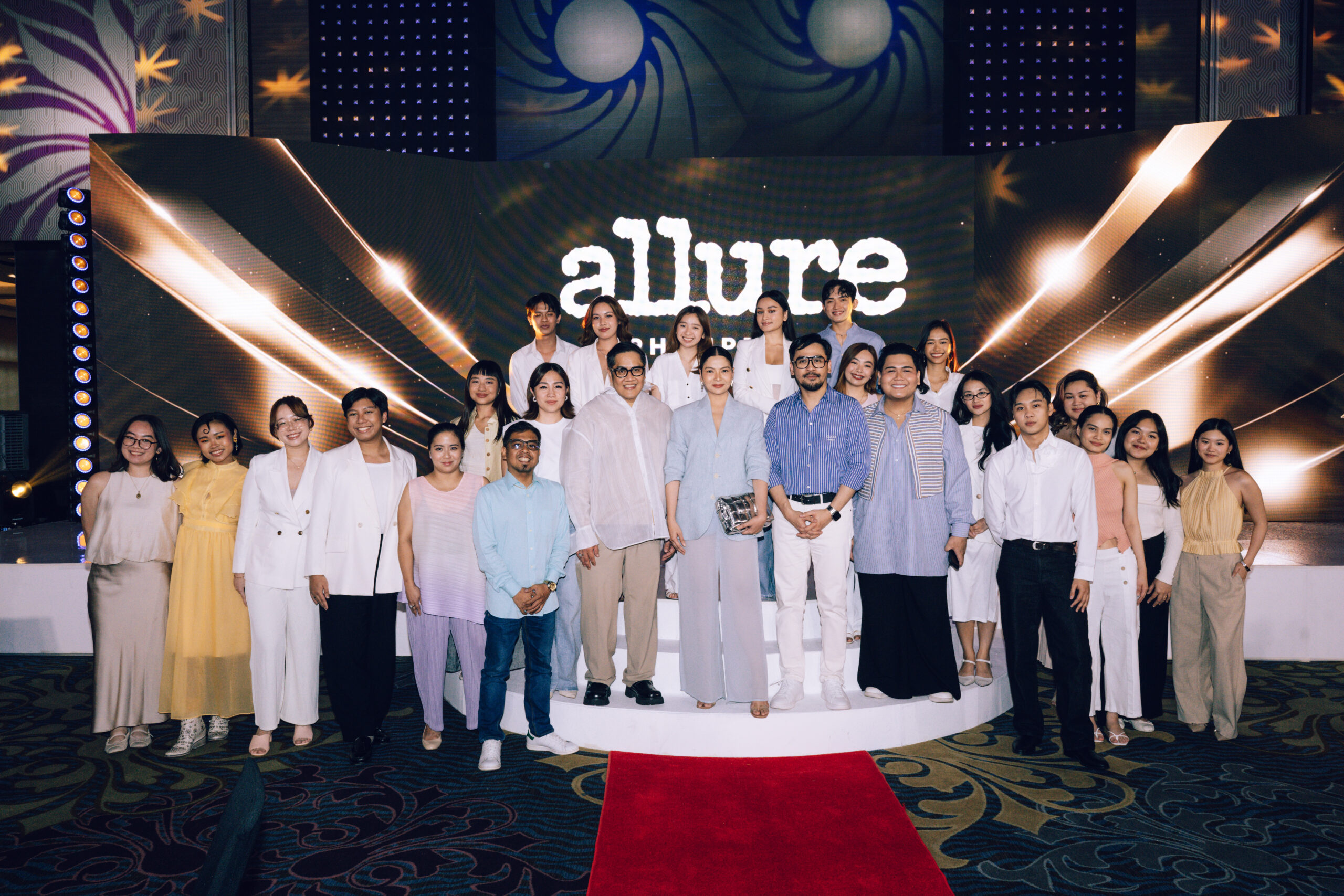 Group of people posing on a stage with a red carpet and a bold backdrop that reads 'allure' behind them.