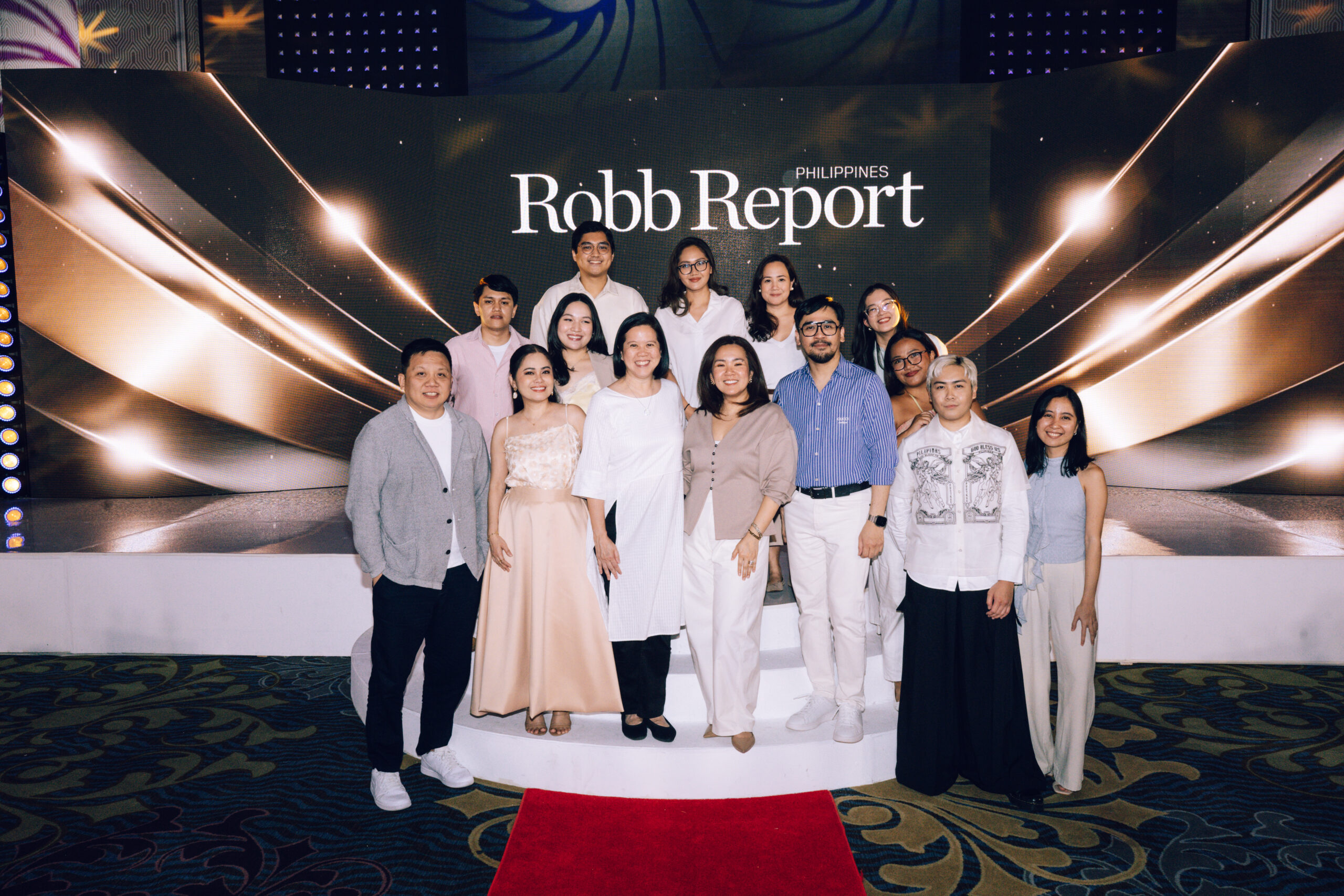 Group of men and women posing on a stage with a large Robb Report Philippines backdrop and golden decorative panels, red carpet in foreground.