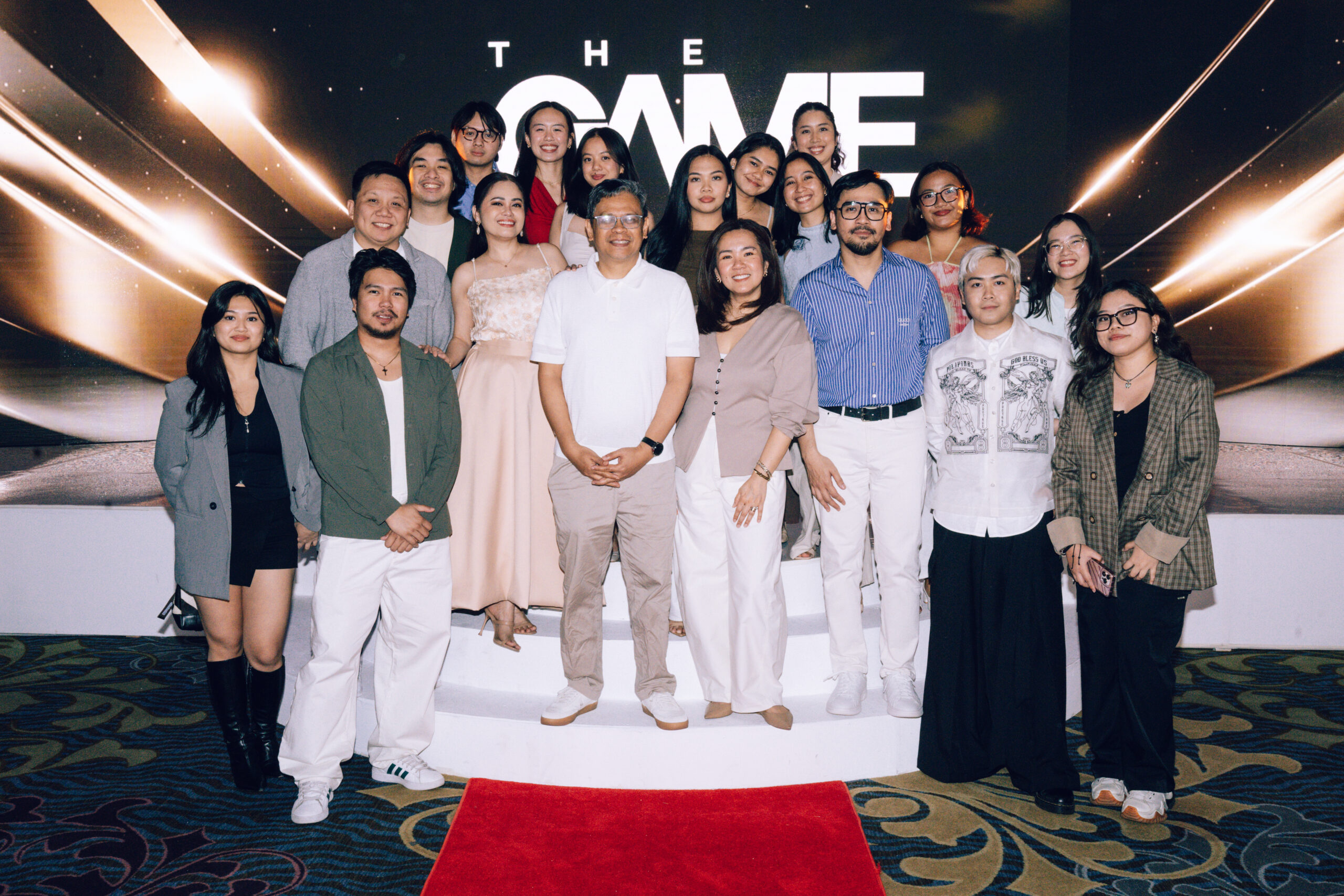 Group of people posing on a stage with a red carpet and a dark backdrop that reads 'THE GAME' behind them.