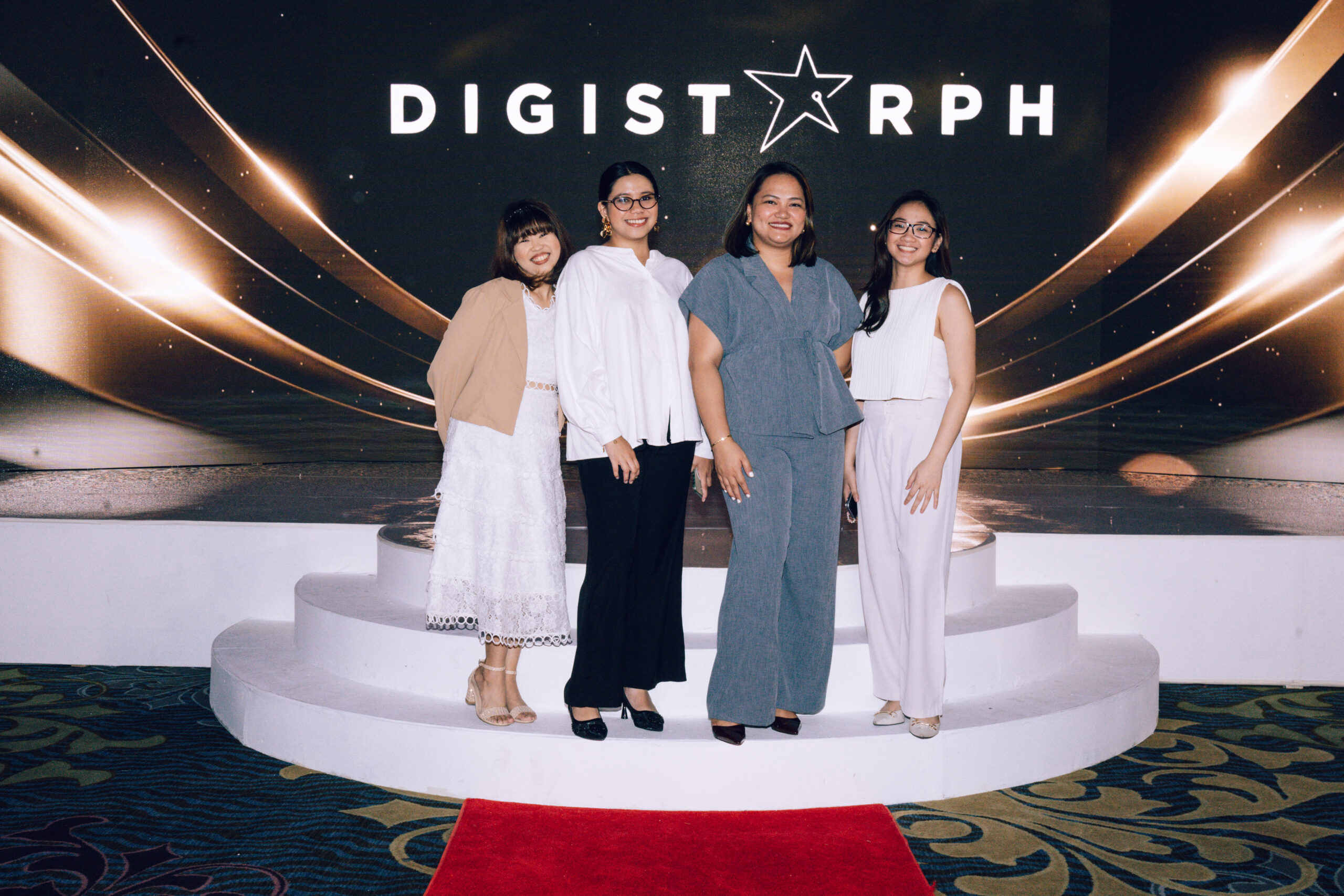 Four women pose on a white circular stage in front of a dark backdrop with gold light streaks and a logo that reads DIGI... on the wall behind them.