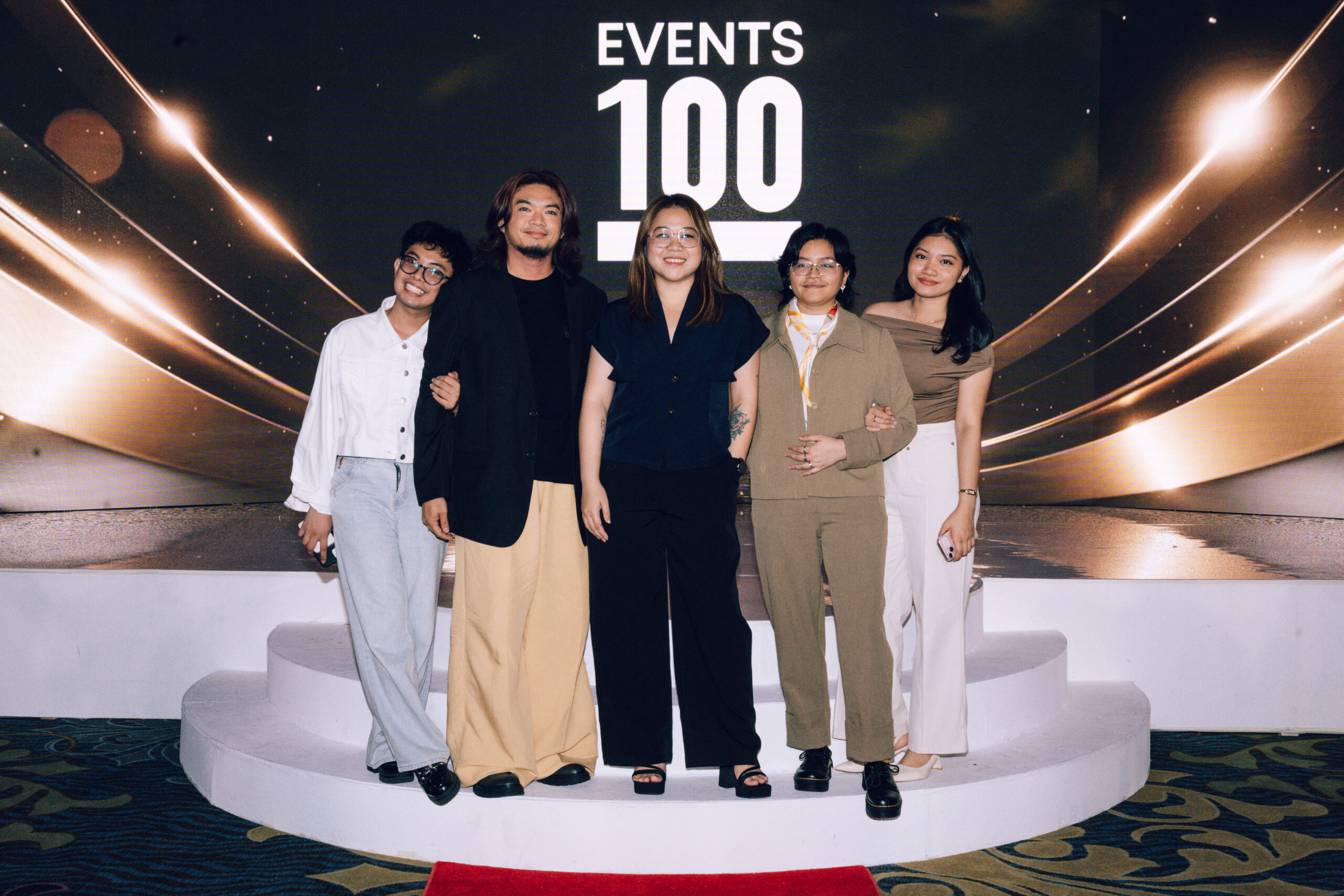 Five people pose on a curved white podium on a stage with a dark background and 'EVENTS 100' visible behind them.