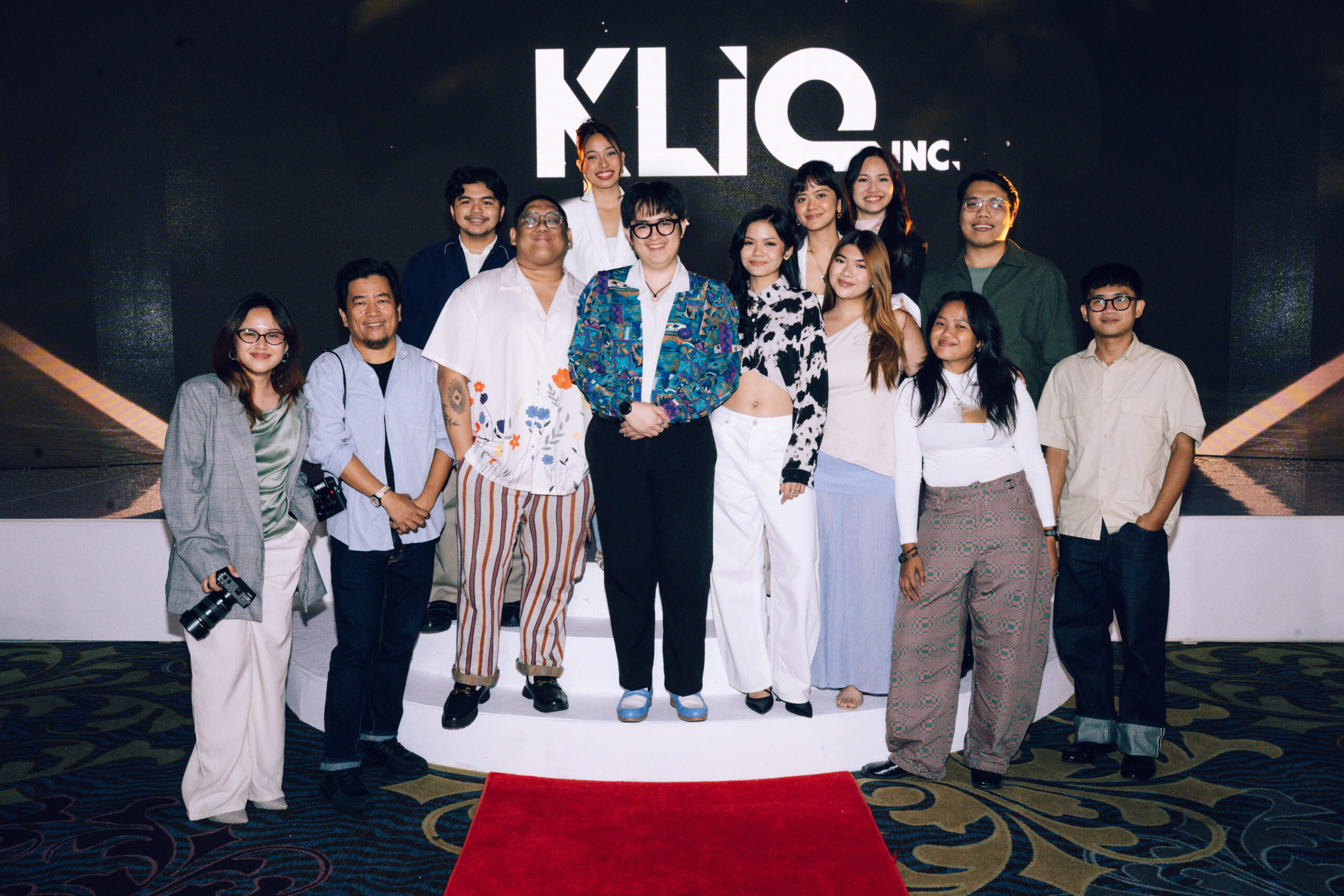 Group of diverse adults posing together on a stage with a large white logo behind them and a red carpet in the foreground, smiling for the camera.