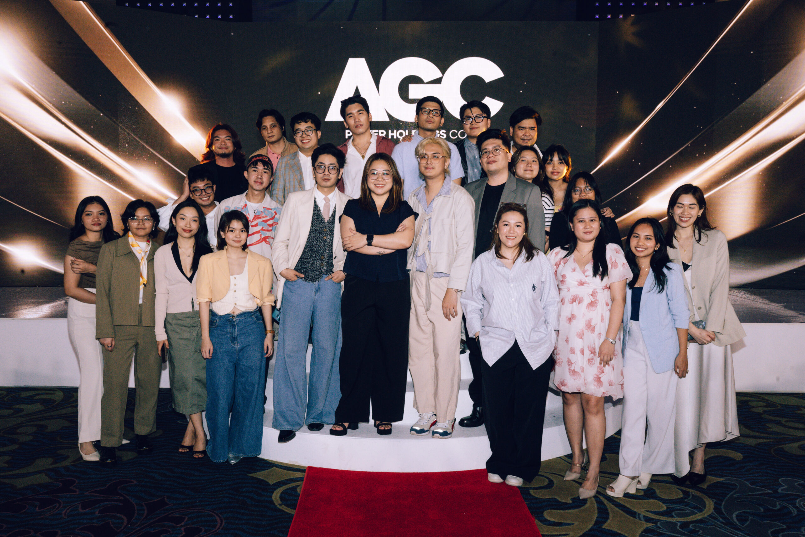 Group of young professionals posing on a stage with a large AGC backdrop, smiling at the camera in smart casual attire.