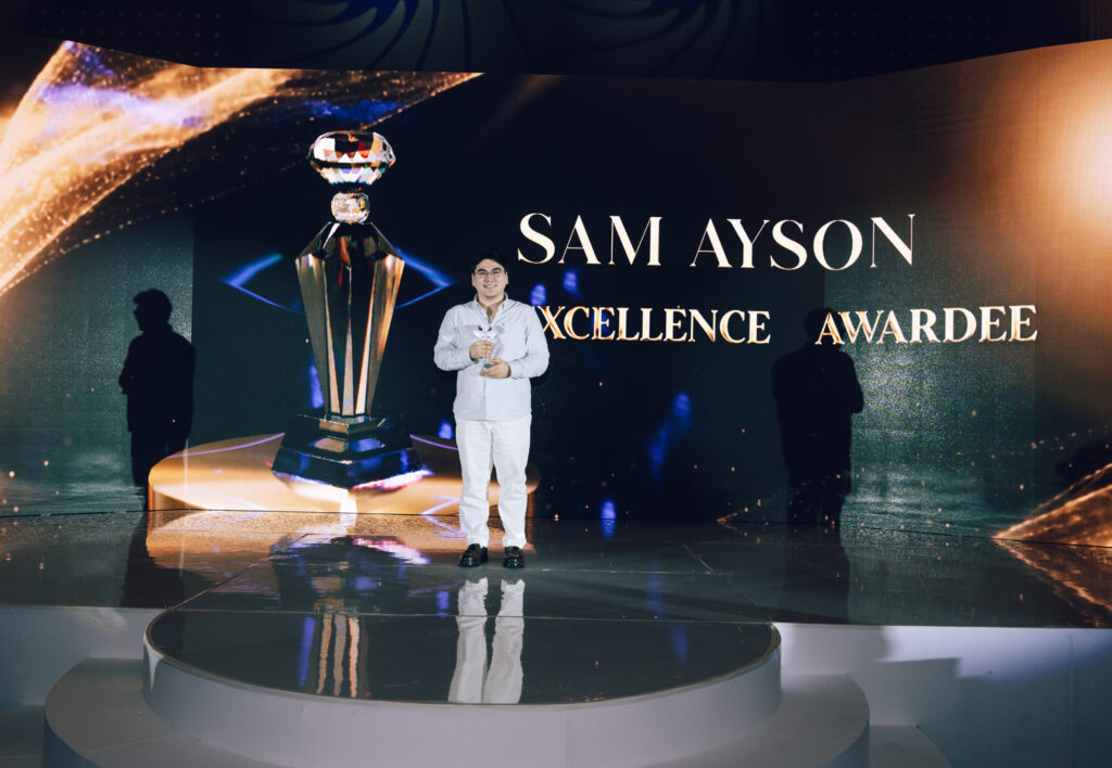 Man in white holding a trophy on a decorated stage, with a large screen showing 'SAM AYSON' and 'EXCELLENCE AWARDEE' in the background.