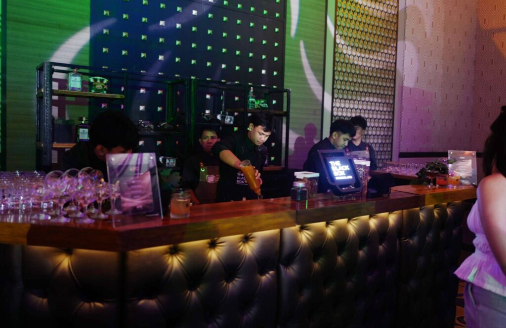 Bartender pours an orange cocktail behind a leather-tufted bar, with bottles on shelves and colorful club lighting in the background.