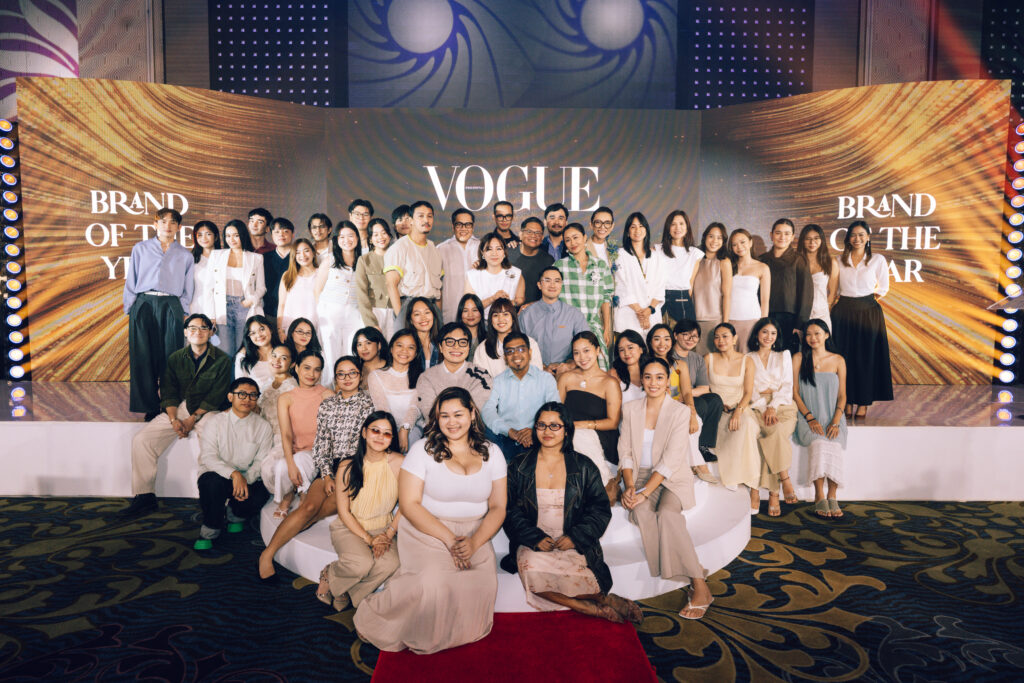 Group of diverse young adults posing on a stage with a Vogue backdrop and orange-bronze panels in the background, posing for a team photo at an event.