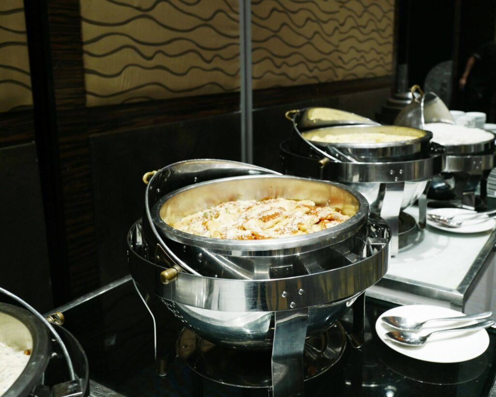 Buffet setup with stainless steel chafing dishes holding hot food; serving spoons and white plates nearby.