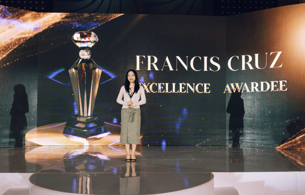Woman standing on a stage holding an award in front of a screen that says 'Francis Cruz Excellence Awardee' with a crystal trophy on display.