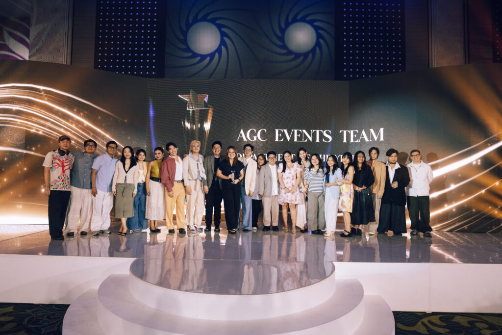 Group photo of AGC Events Team on stage, posing together in front of a illuminated backdrop with 'AGC EVENTS TEAM' text