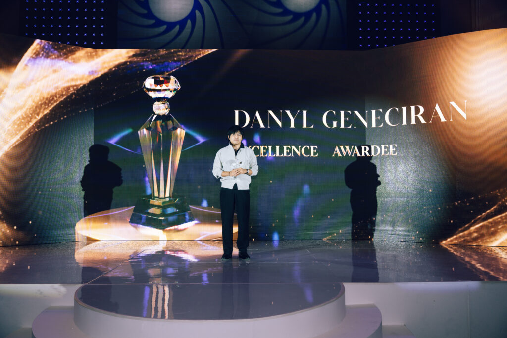 Man in a light shirt stands on a glossy stage in front of a large screen showing a crystal trophy and the words 'Excellence Awardee' in a formal event setting