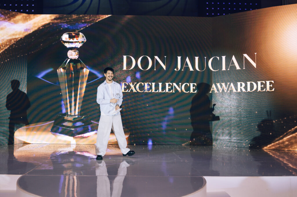 Man in light clothing holding an award on a stage in front of a large screen that reads 'DON JAUCIAN EXCELLENCE AWARDEE' with a crystal trophy graphic beside him