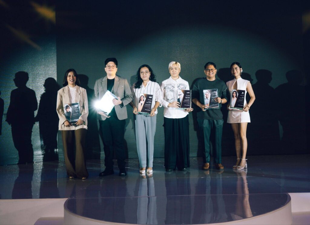 Six individuals stand on a stage, each holding an award booklet or magazine and posing for a photo at an event.