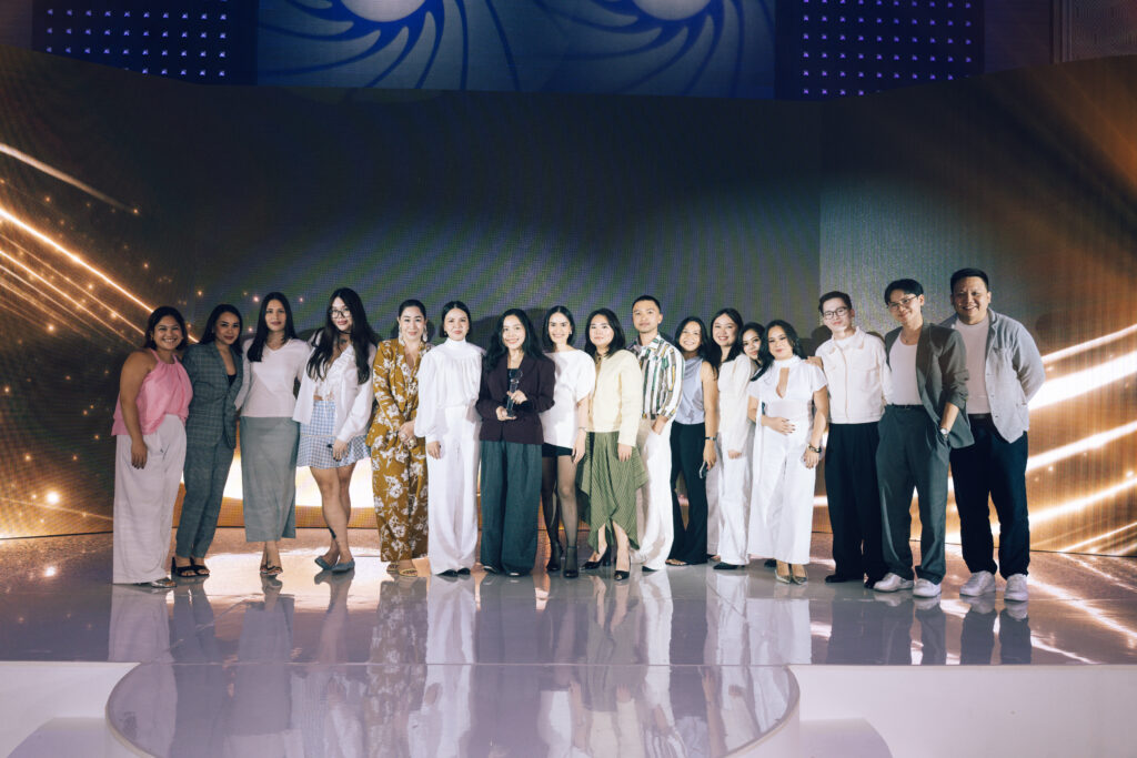 Group of about twenty people posing on a lit stage, some in formal wear, with a curved reflective floor and a digital backdrop.