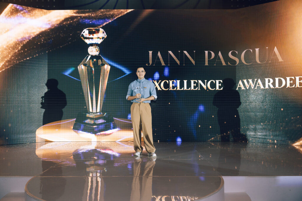 Man on stage holding a crystal trophy during an Excellence Award ceremony, with a large trophy image and EXCELLENCE text in the backdrop.