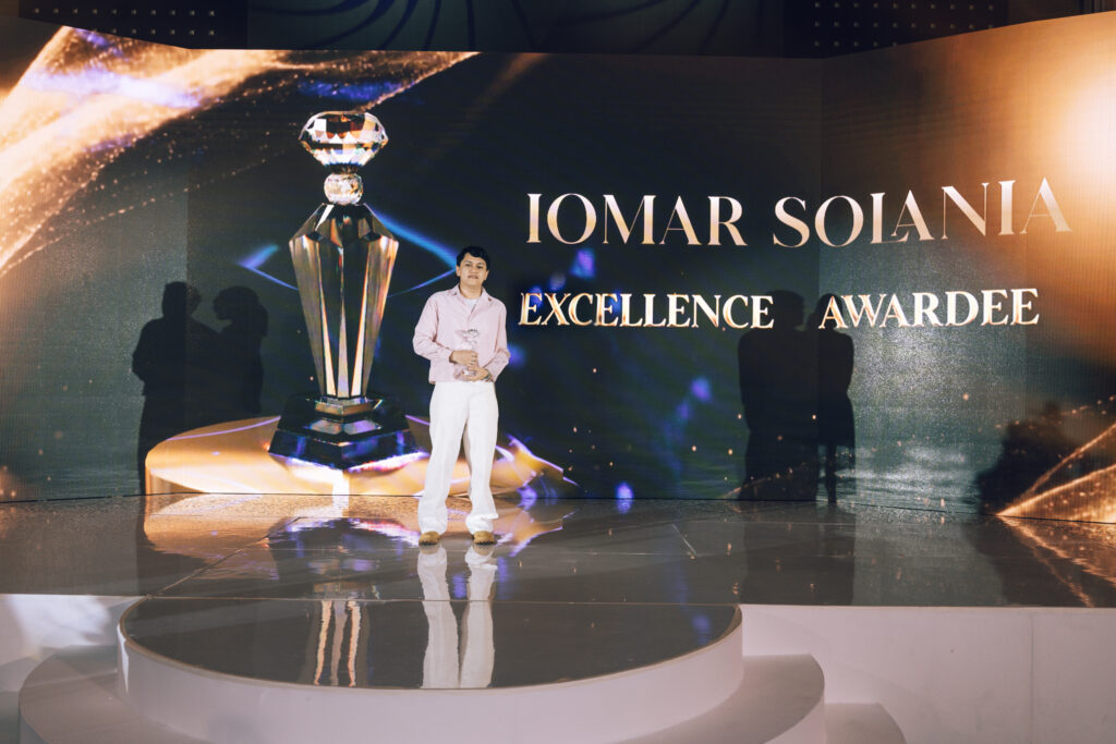 Young man posing on a glossy stage in front of a backdrop showing a crystal trophy and the text 'EXCELLENCE AWARDEE'.