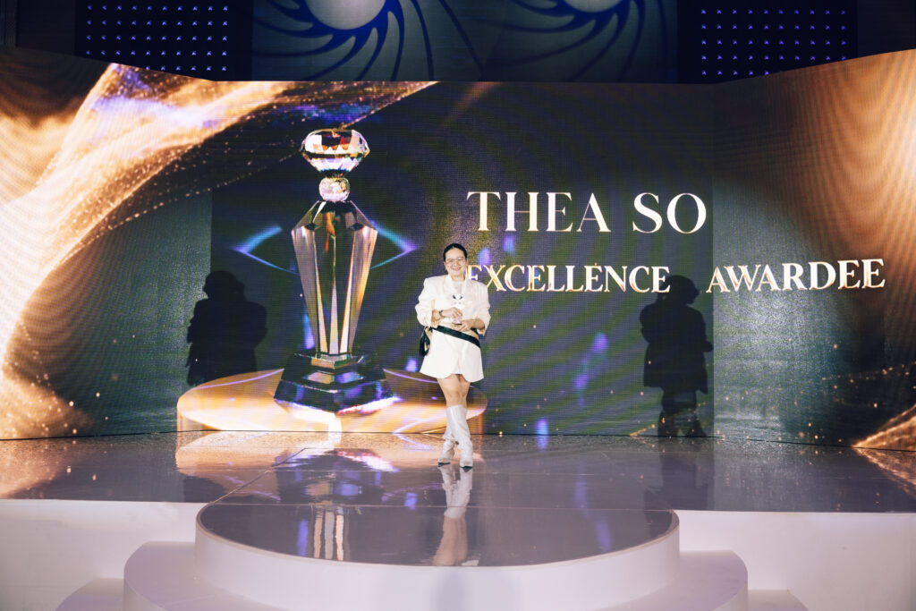 Woman in a white outfit stands on a curved stage, holding a crystal trophy as a large 'Excellence Awardee' backdrop glows behind her.