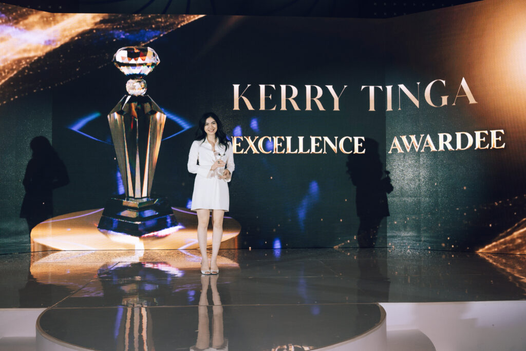 Woman in a white dress stands on a stage, smiling, holding a glass award with a large trophy graphic and 'KERRY TINGA EXCELLENCE AWARDEE' backdrop behind her.