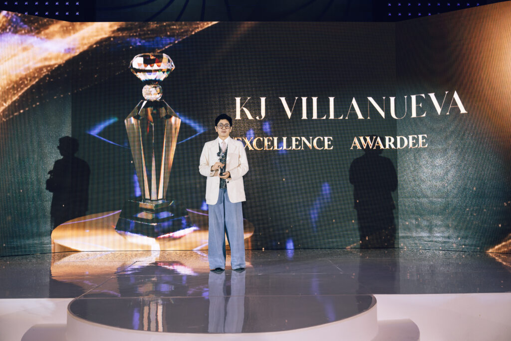 Man in a white blazer holding a crystal trophy on a stage, with a large screen showing 'KJ VILLANUEVA EXCELLENCE AWARDEE' behind him.