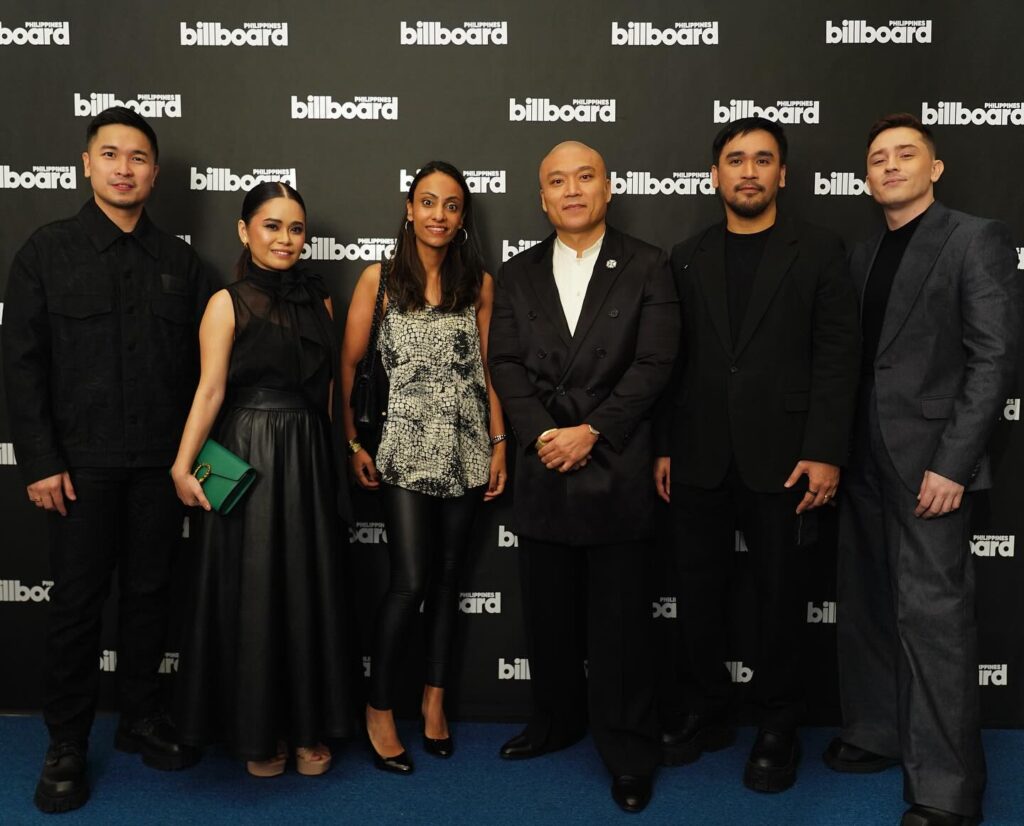 Six people posing on a blue carpet in front of a black Billboard backdrop, all wearing black outfits.