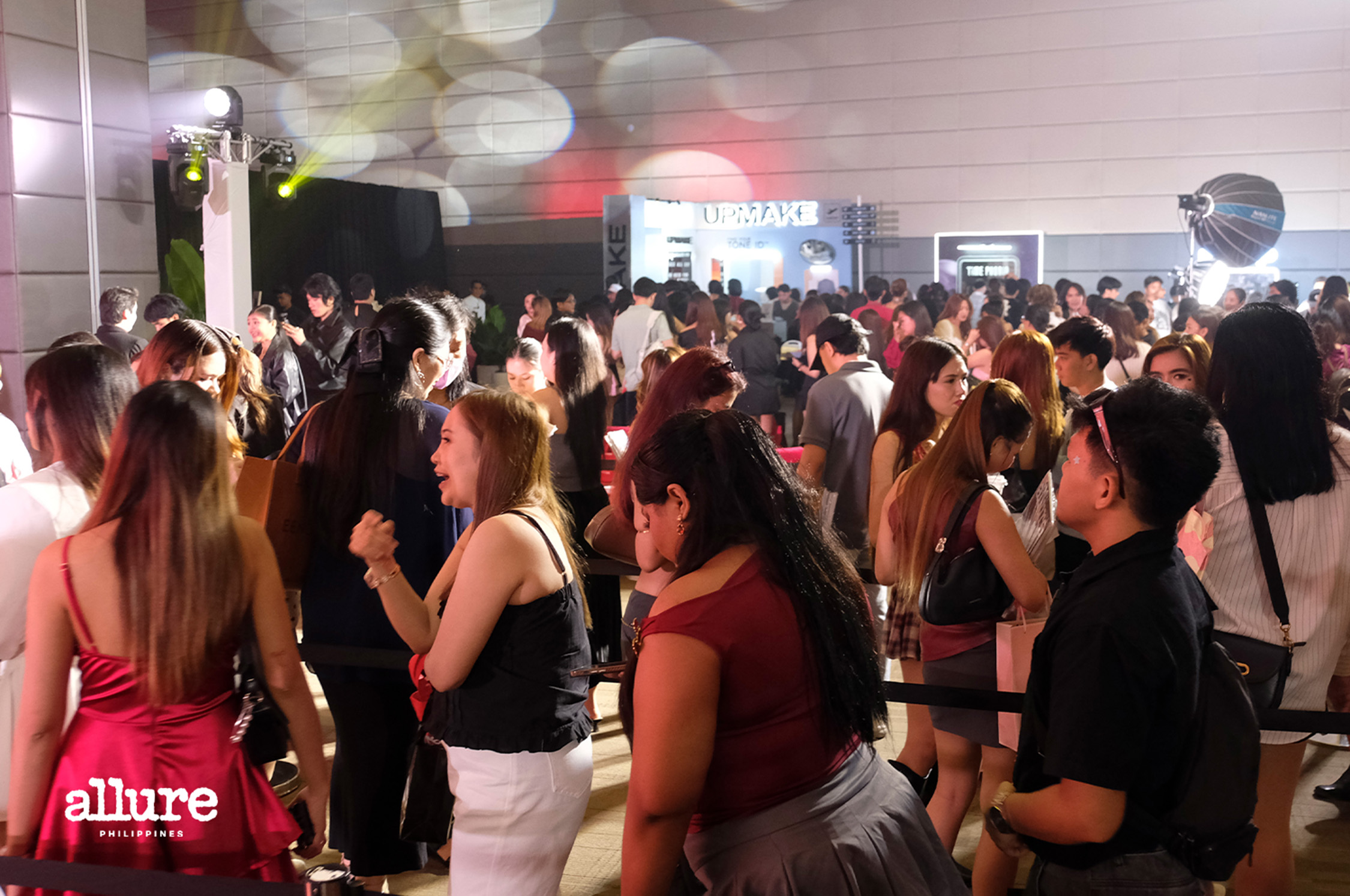 Crowded expo or fashion event with attendees chatting; foreground shows a woman in red with a tote that says 'allure PH' on her bag.