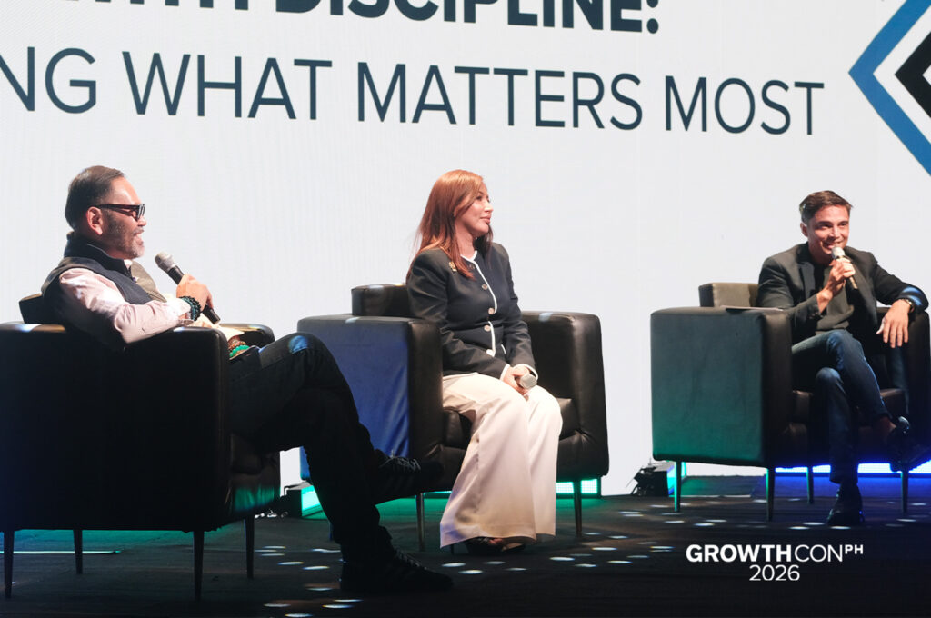 Three panelists seated in black chairs on a lit stage during GrowthCON PH 2026, each holding a microphone.