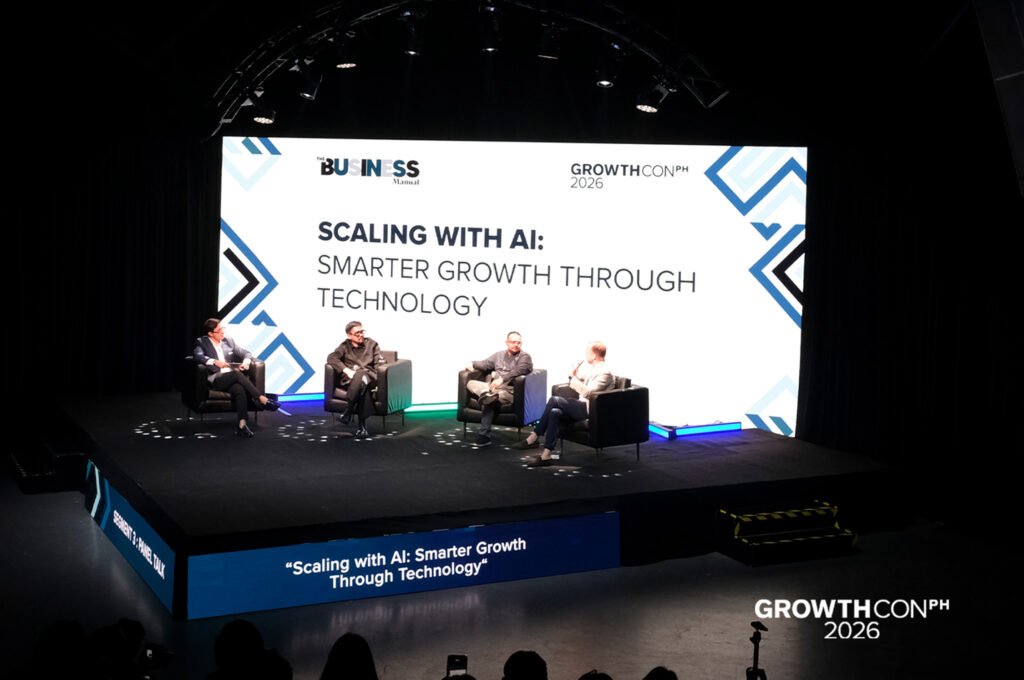 Panel of five speakers seated in black chairs on a stage, large illuminated screen behind them displaying the title 'SCALING WITH AI: SMARTER GROWTH THROUGH TECHNOLOGY' at GrowthCon 2026.