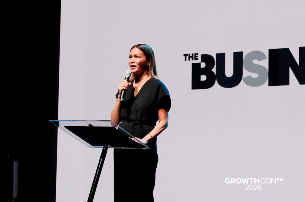 Woman in a black outfit speaks into a handheld microphone at a clear podium on a stage, GrowthCon backdrop visible.
